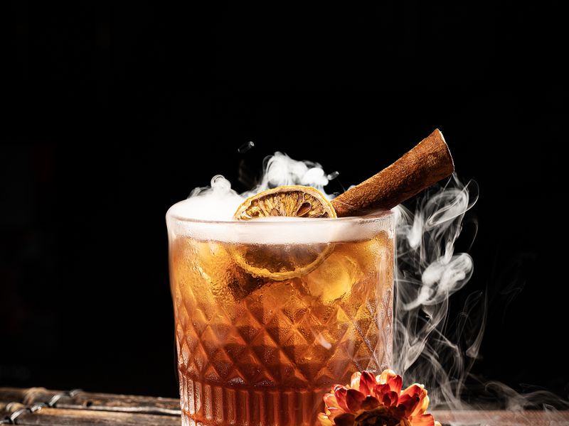 Smoke curling off a crystal glass of bourbon with an orange slice and cinnamon stick